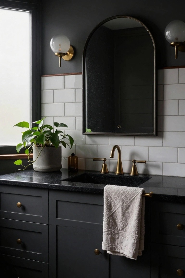 Bathroom vanity featuring a black granite countertop and undermount sink, flanked by dark cabinets with brass knobs, a tall arched mirror with black frame, two brass wall sconces with white globe shades, white subway tile backsplash with terracotta trim, a potted green plant, a small glass bottle, and a white towel on a brass bar.