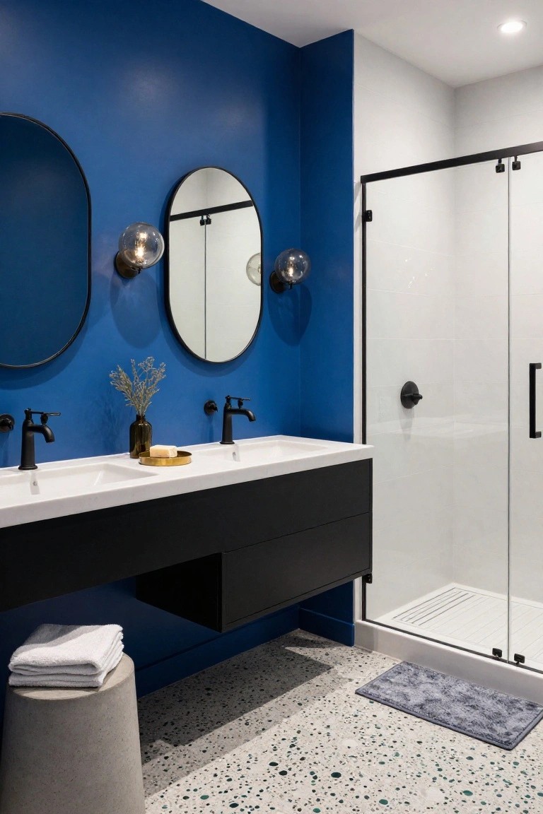 Contemporary bathroom with deep blue accent wall, two oval black-framed mirrors flanked by black globe wall sconces, floating black vanity with double white sinks and matte black faucets, glass shower enclosure, concrete stool with towels, and terrazzo floor.