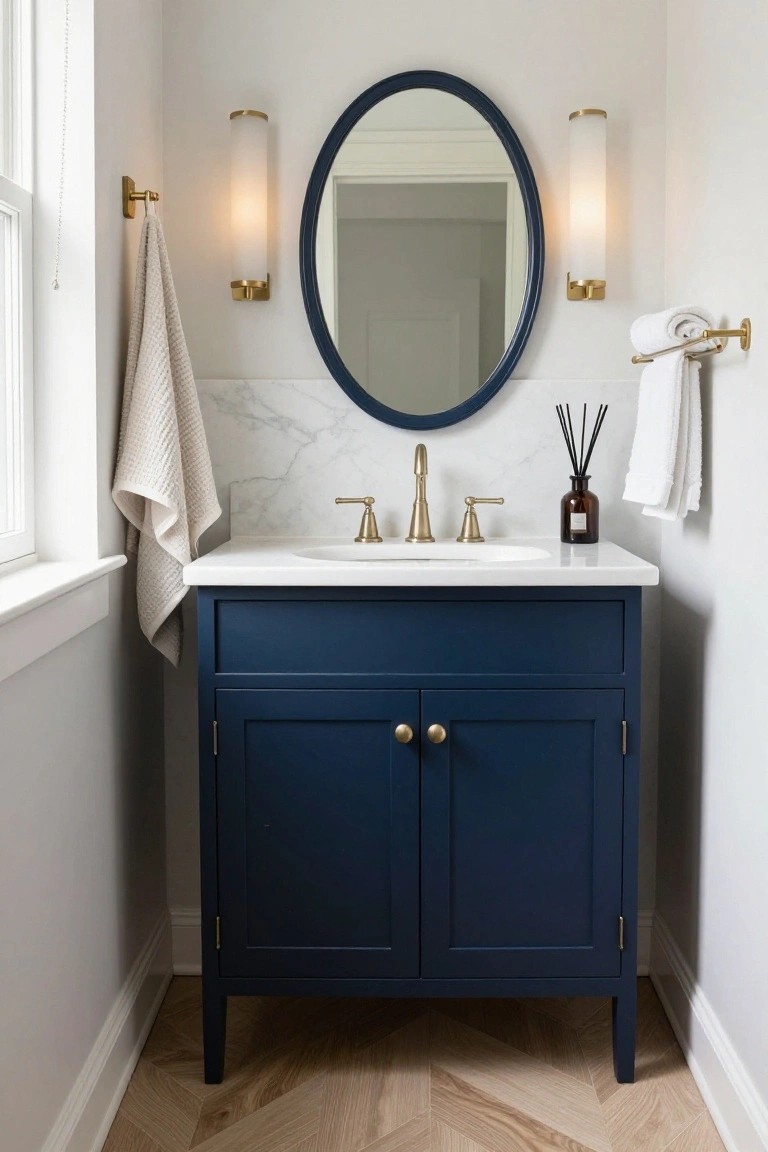 Small bathroom vanity with navy blue cabinet, white marble countertop, gold bridge faucet, oval blue-framed mirror centered above flanked by two gold wall sconces with white cylindrical shades, hanging white towels, reed diffuser, and herringbone wood floor.
