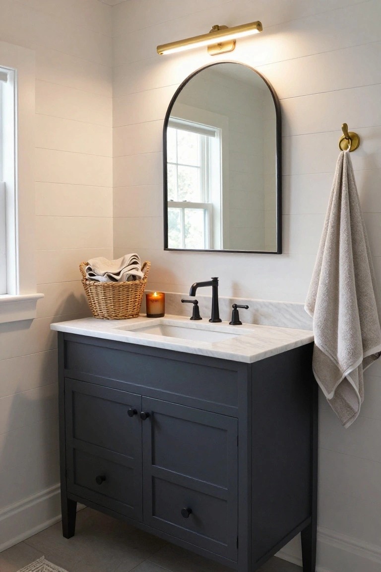 Bathroom vanity setup featuring a navy cabinet with white marble top, undermount sink, black faucet, arched black-framed mirror, gold arched wall sconce, wicker basket of towels, hanging towel, and shiplap walls.