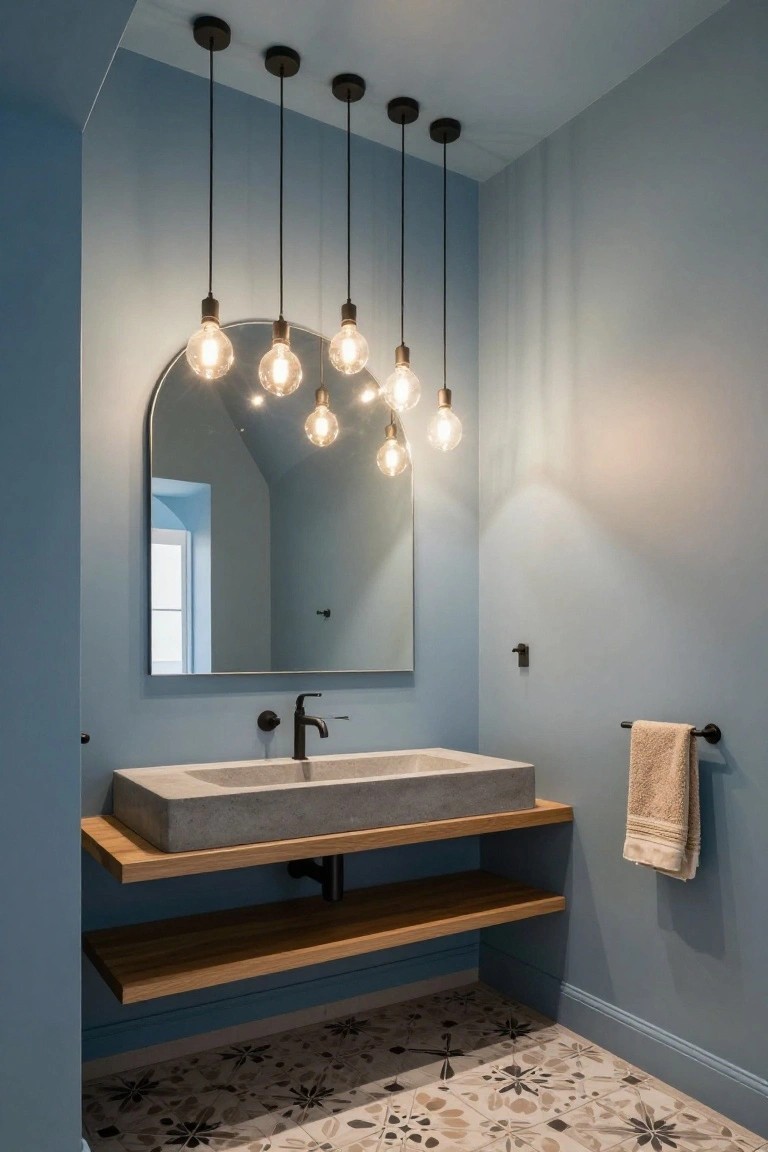 A light blue vaulted ceiling bathroom with five black pendant lights featuring exposed bulbs hanging above a concrete vessel sink on a wooden shelf, an arched mirror, black faucet, towel bar, and patterned tile floor.