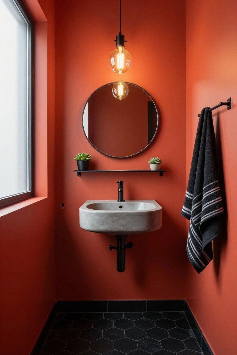Compact powder room featuring orange walls, black hexagonal floor tiles, wall-mounted rectangular concrete sink with black faucet, round black-framed mirror with two potted succulents on a black shelf, hanging exposed-bulb pendant light, black towel rail with striped towel, and a window.