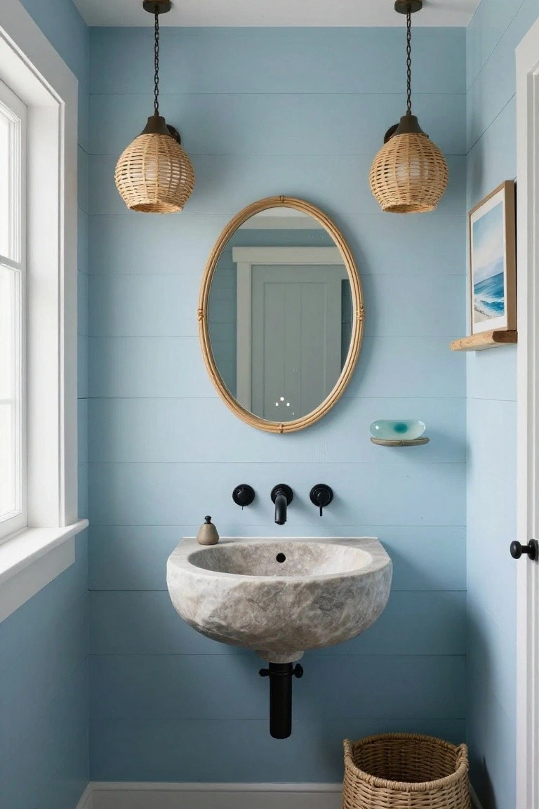 Small powder room with light blue shiplap walls, hanging rattan pendant lights over a white marble vessel sink with black faucets, oval rattan mirror, glass shelf, and woven basket on floor.