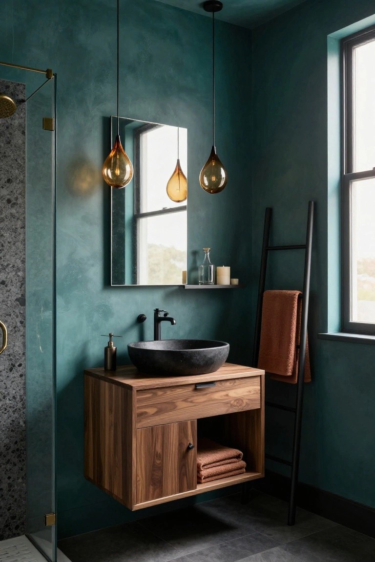 Teal textured bathroom with floating walnut vanity, black stone sink, brass shower enclosure, black ladder towel rack holding orange towels, and three amber glass teardrop pendant lights hanging from the ceiling.