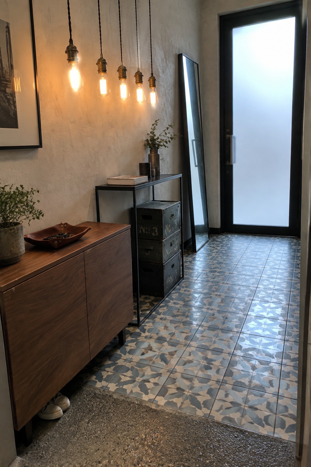 An interior entryway featuring a row of exposed bulb pendant lights above a wooden console, stacked metal crates, a tall mirror, and a black door with patterned tile flooring.