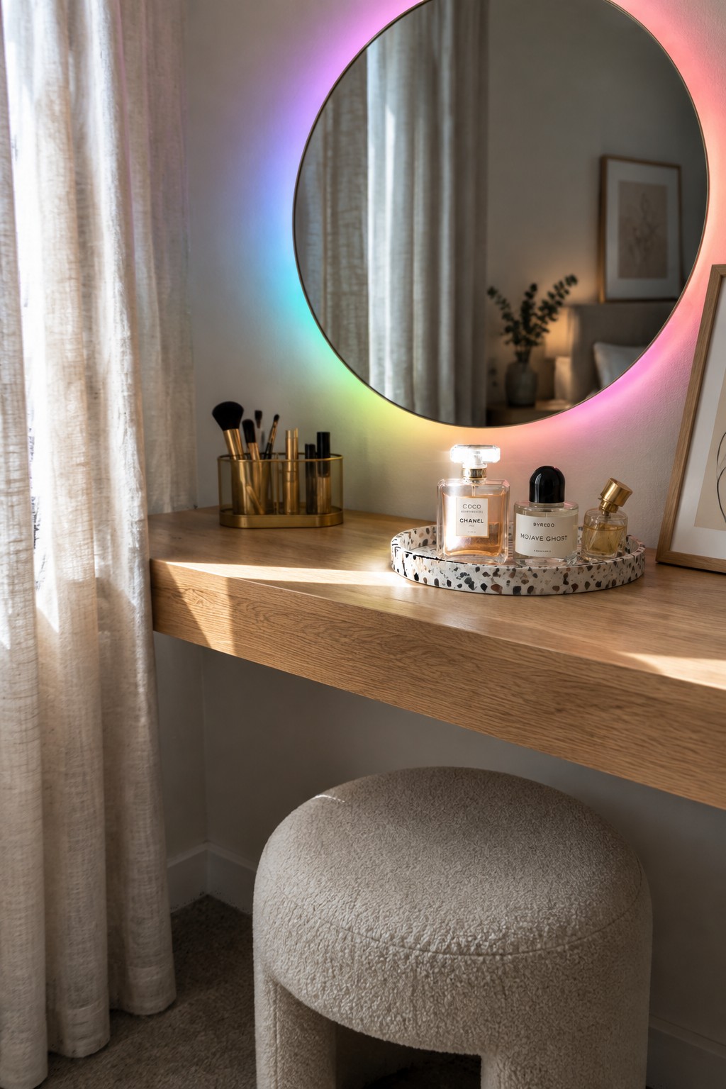 A round mirror with a glowing multicolored LED ring mounted above a light wood vanity, with perfume bottles on a terrazzo tray and a makeup brush holder nearby.