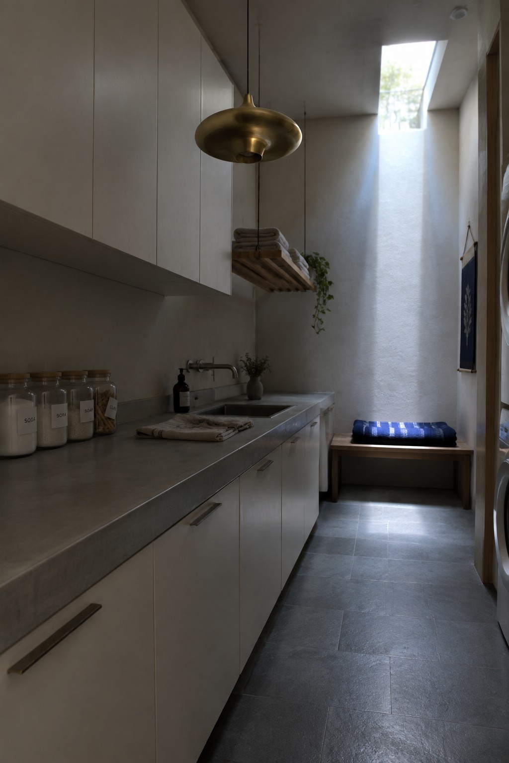 A narrow laundry room with white cabinets, a gray concrete countertop, a large brass pendant light hanging above the sink, and a skylight at the far end.