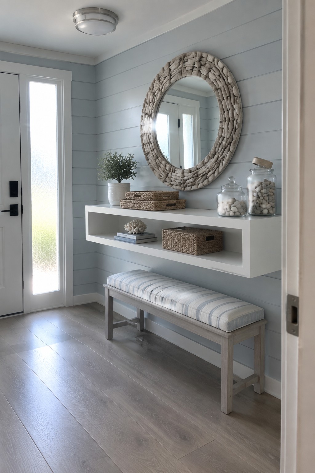 A foyer with light blue shiplap walls, a white floating shelf holding baskets and books, a striped bench, a round stone-framed mirror, and a flush mount ceiling light.