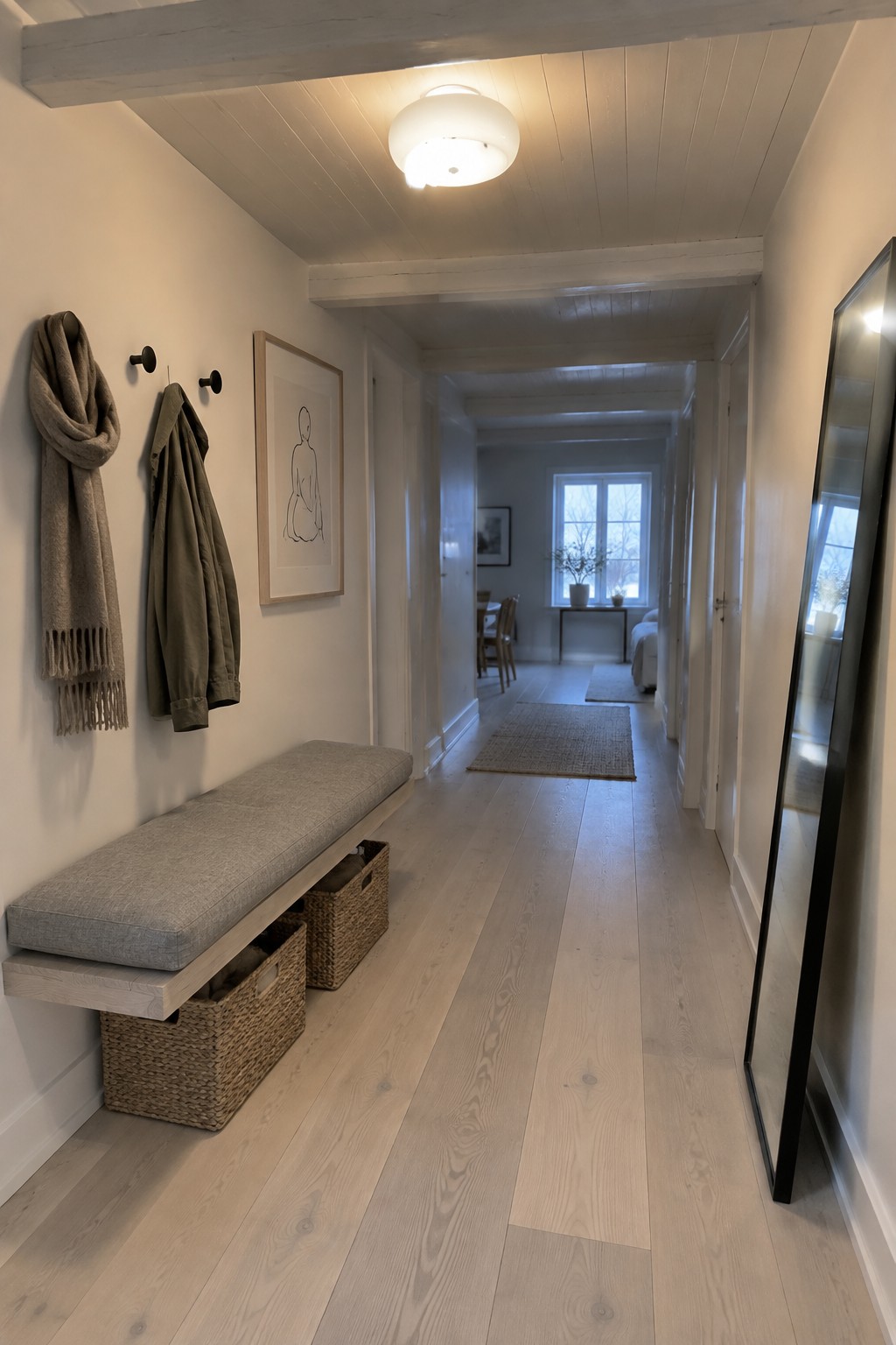 A narrow interior hallway with light wood flooring, a wall-mounted bench with woven storage baskets, coats on hooks, framed art, and a round white flush mount ceiling light.