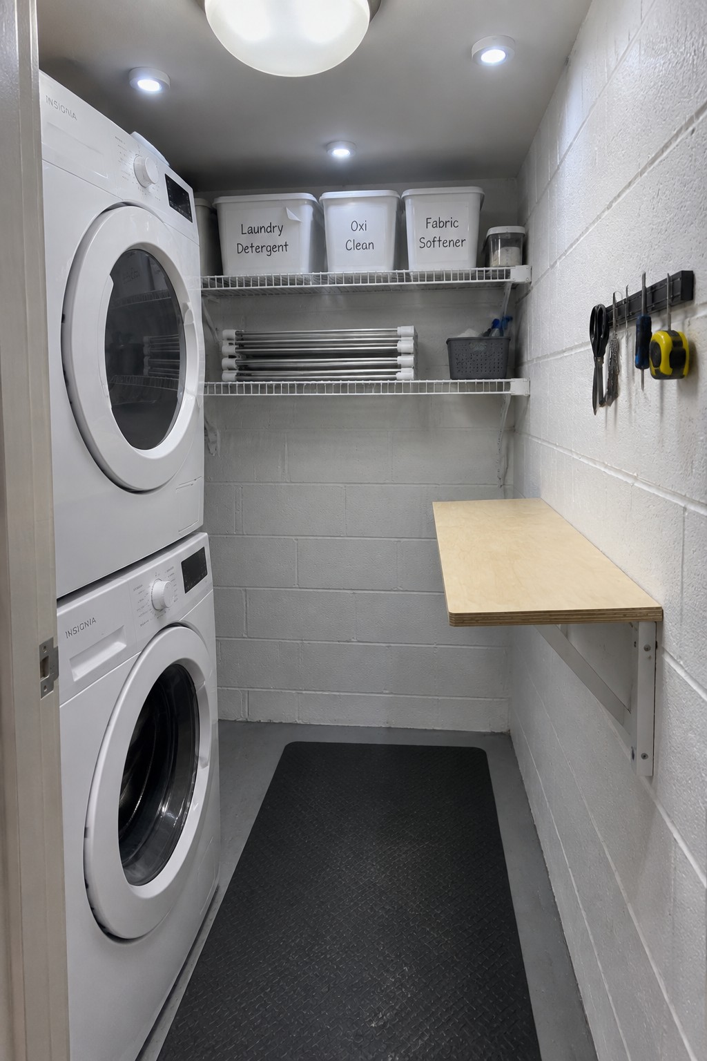 Laundry room interior with stacked white washer and dryer on the left, wire shelves with labeled bins, a wooden fold-down table on the right wall, and multiple ceiling light fixtures including one large globe and smaller recessed lights.