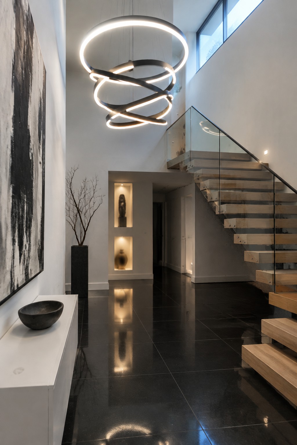 A modern two-story foyer with a large multi-ring chandelier hanging from the ceiling above a glossy black floor and a floating wood staircase with glass railing.