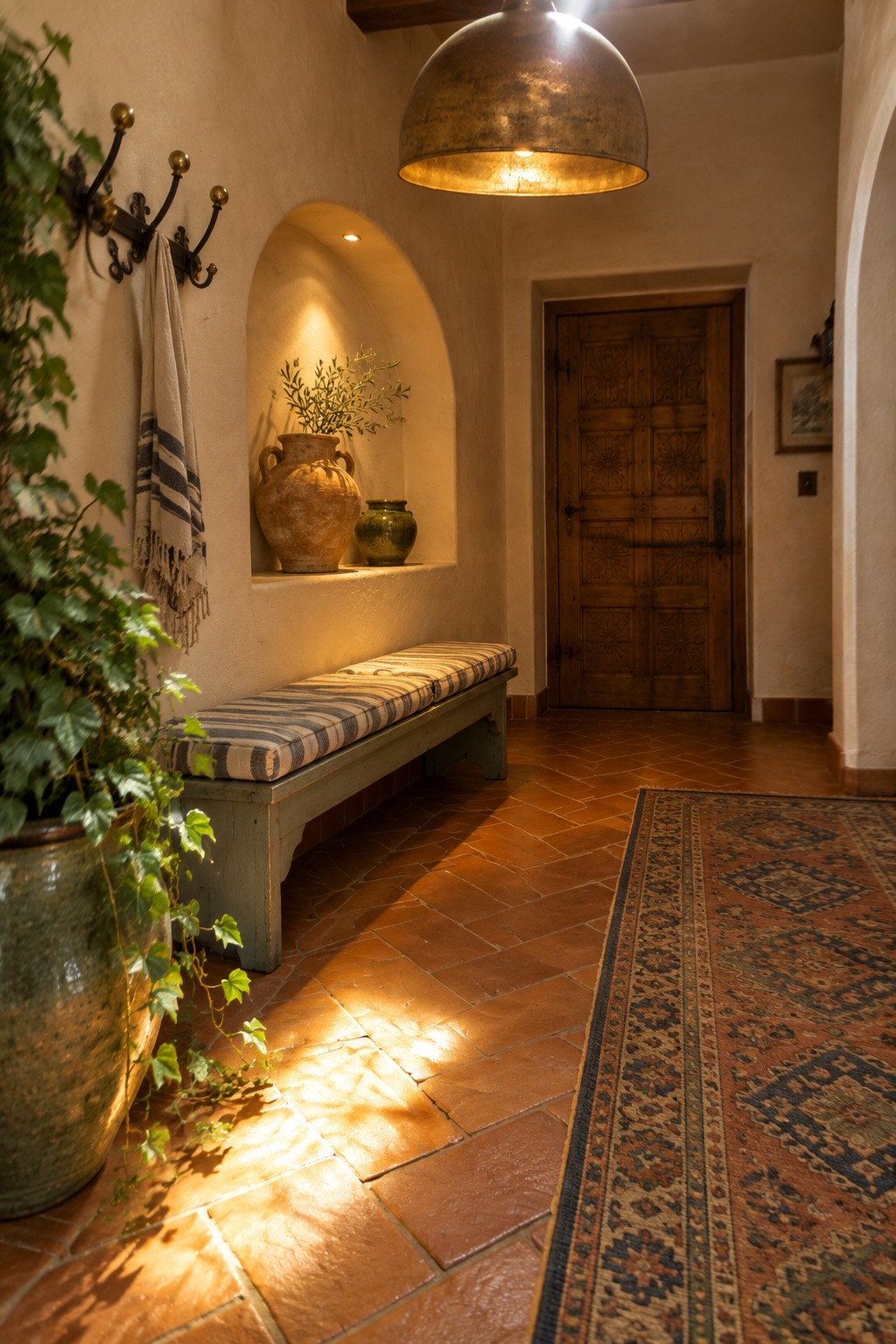 An interior entryway with a large brass pendant light hanging above terracotta tile flooring, a wooden bench, and an arched niche holding pottery.