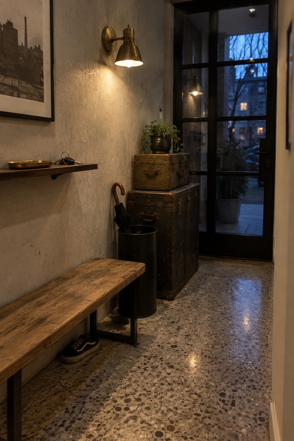 A brass wall sconce mounted above stacked trunks and a wooden bench in a narrow entryway next to a black-framed glass door.