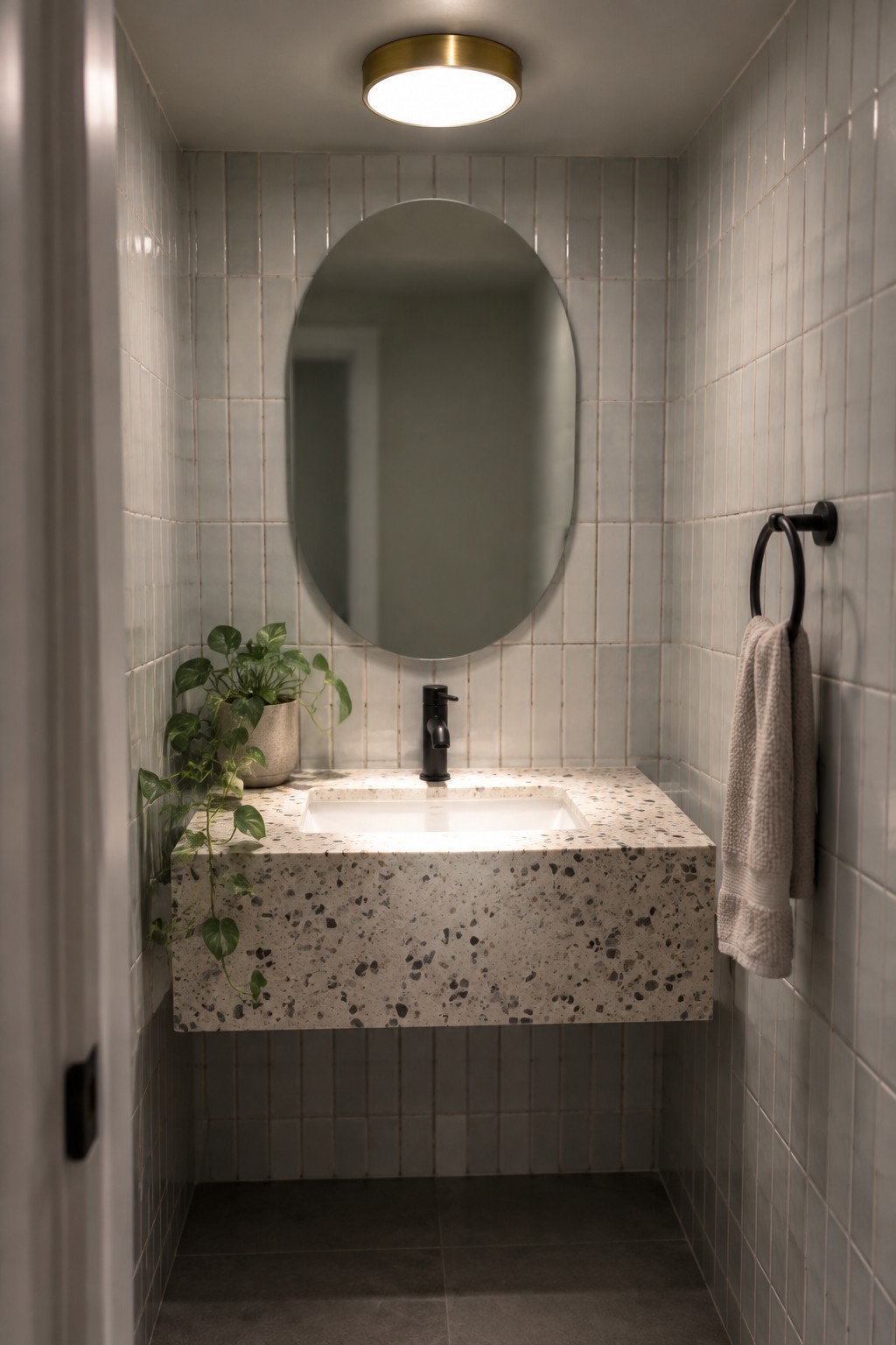 A narrow modern bathroom with light gray wall tiles, a floating speckled terrazzo vanity, round mirror, black faucet, potted plant, and round brass flush mount ceiling light.