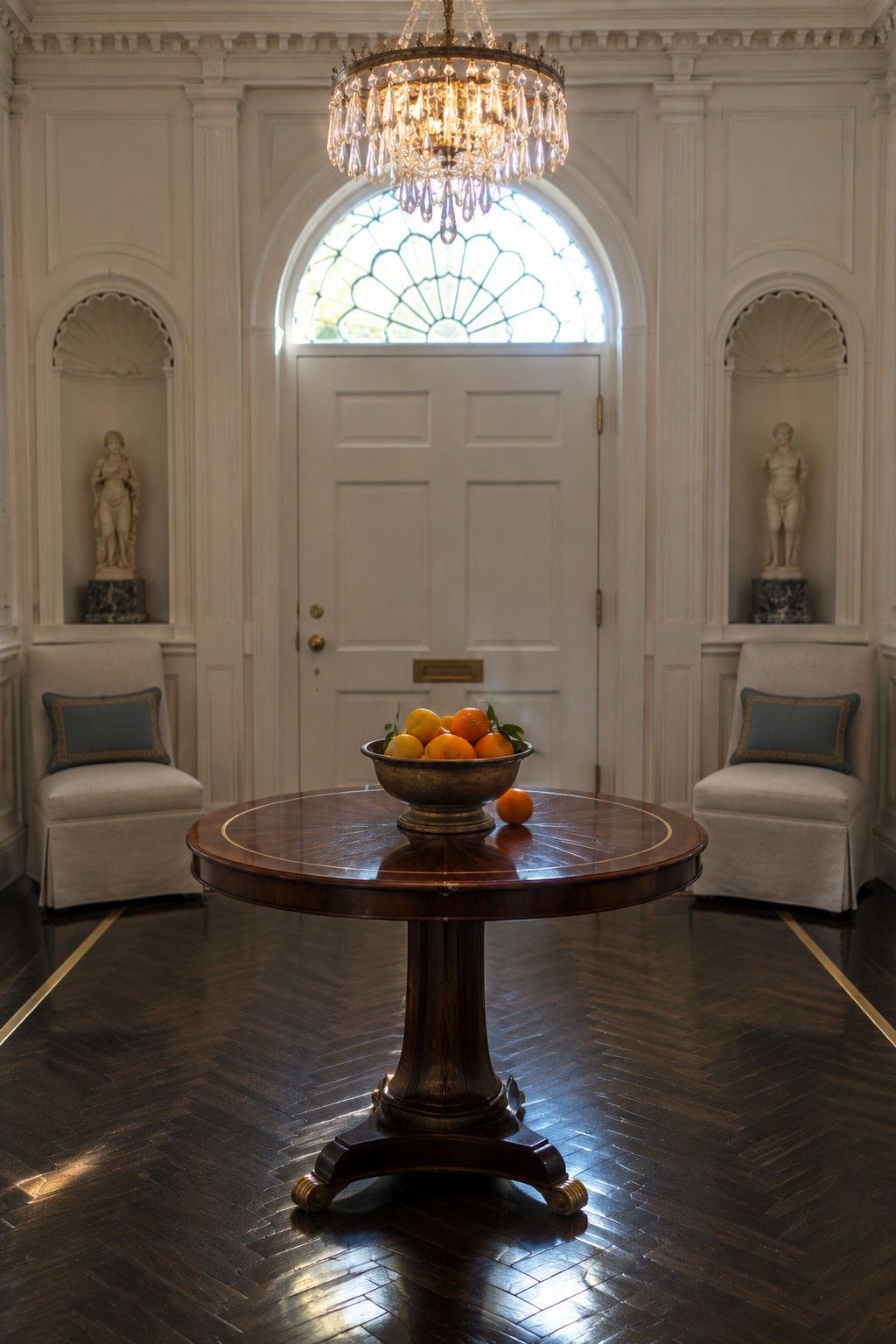 A large crystal chandelier hangs above a round wooden table holding a bowl of fruit in a white traditional foyer with two upholstered armchairs and statues in wall niches.