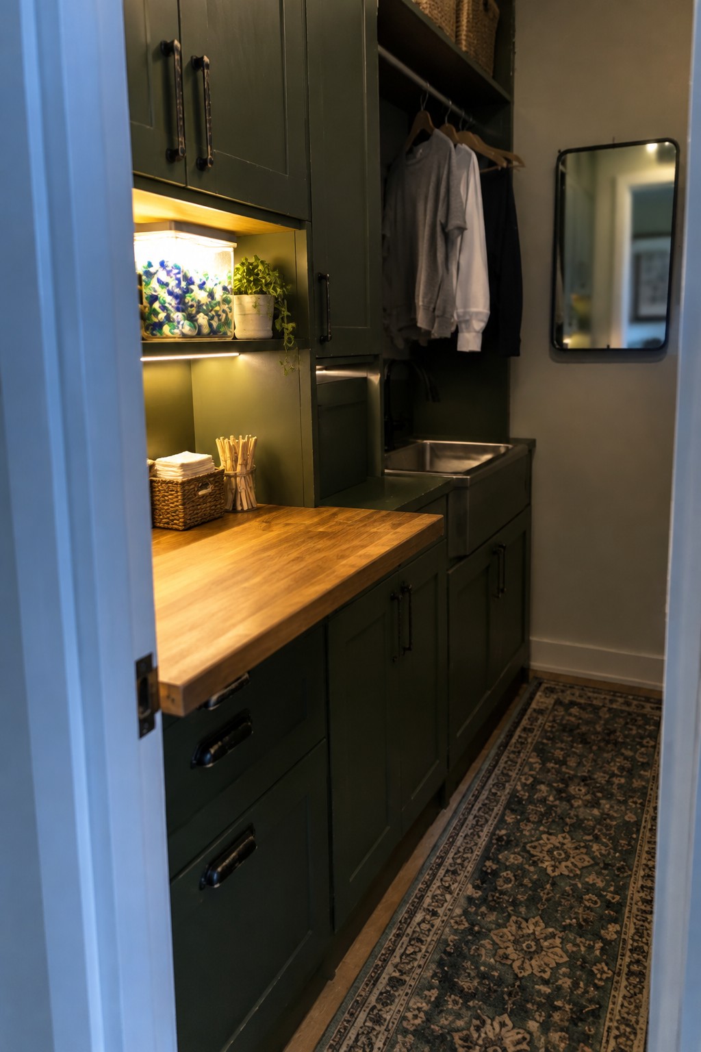 A narrow laundry room with dark green cabinets, a wooden countertop, a stainless sink, and under-cabinet lighting illuminating an open shelf with towels and a plant.