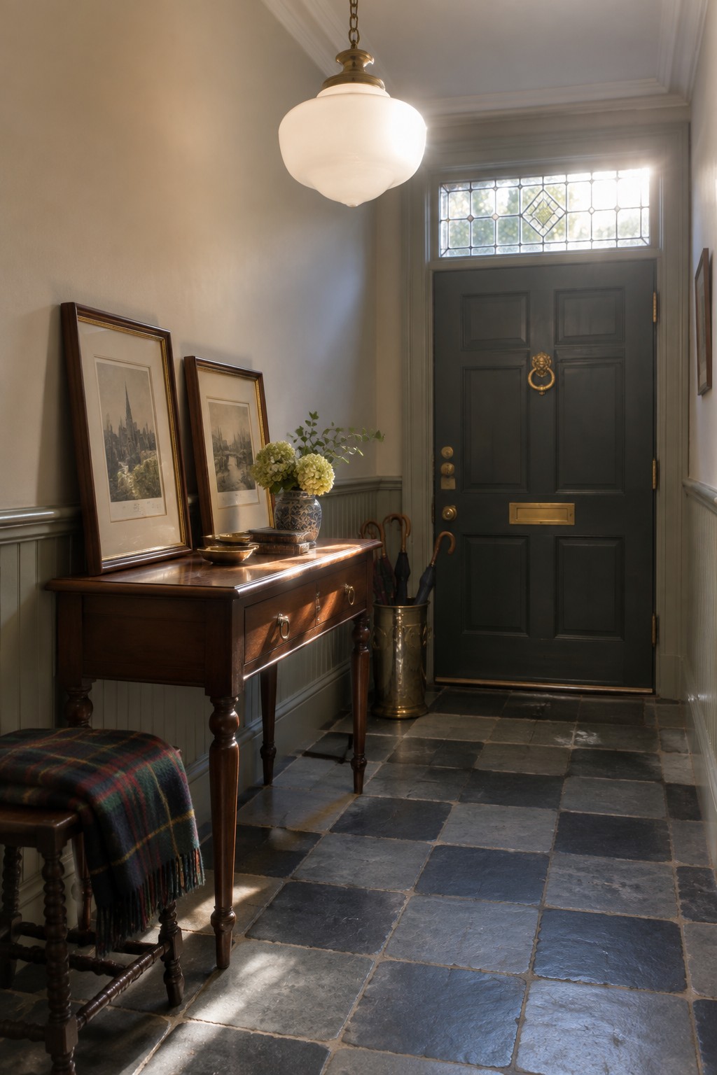 An interior entryway featuring a large white globe pendant light hanging above a dark wood console table, with framed art, a plaid stool, and a dark paneled front door with a stained glass transom.