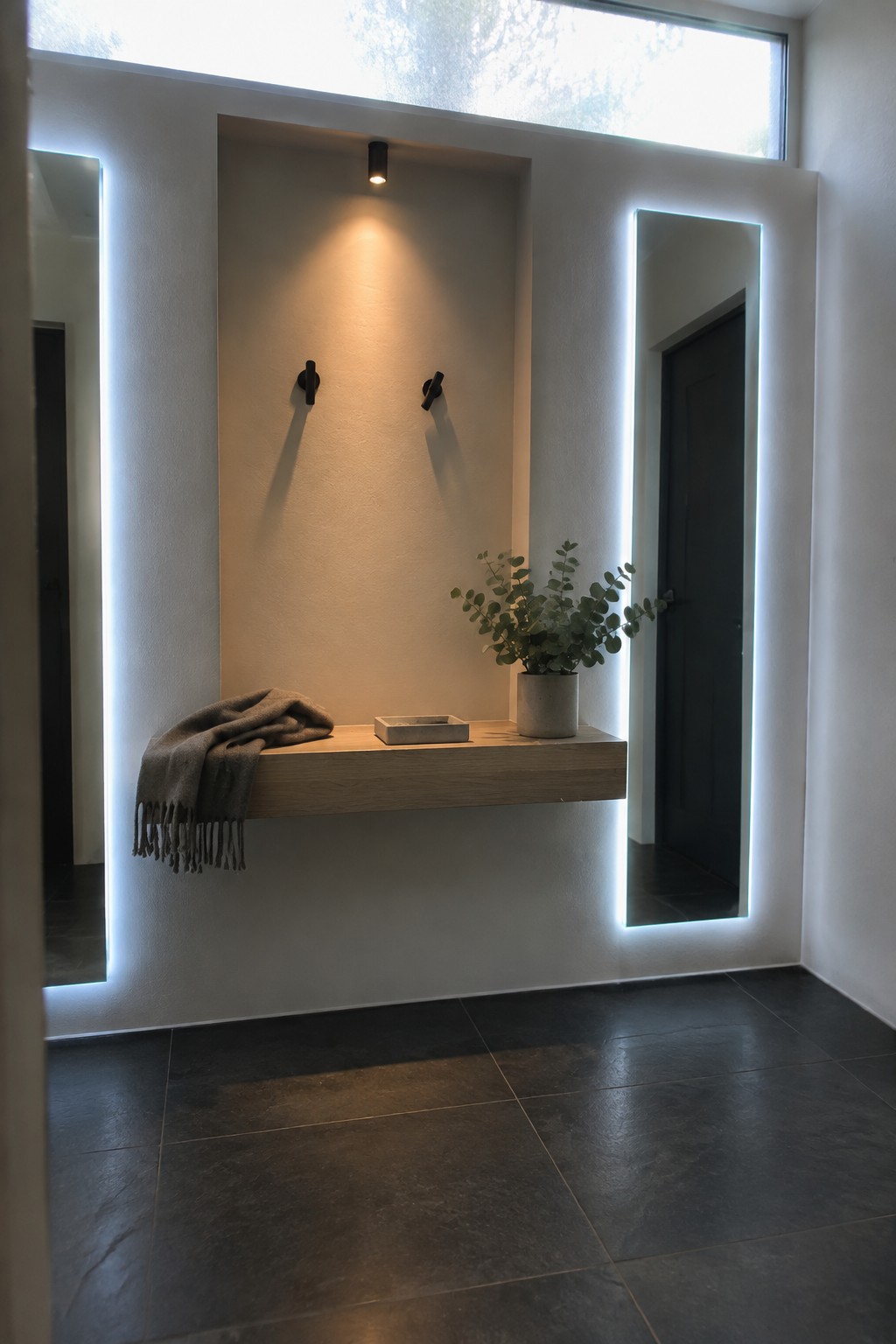 A minimalist interior entryway shows a recessed niche with a light wood floating shelf holding a folded blanket and small tray, flanked by two tall mirrors with bright white LED borders and a single ceiling spotlight above.