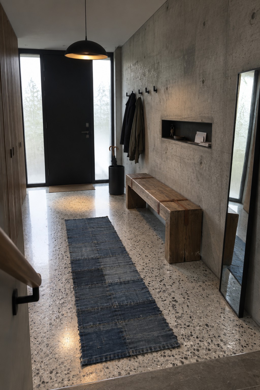 A minimalist interior foyer featuring a black dome pendant light, a wooden bench against a concrete wall, a blue patchwork runner on terrazzo flooring, and a black front door with glass panels.