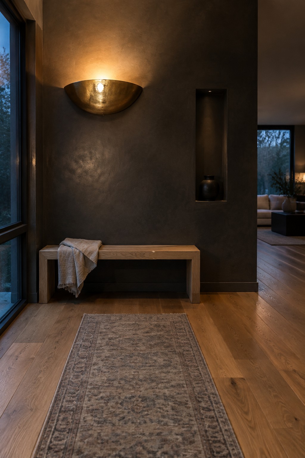 A minimalist interior entry with a light wood bench against a dark textured wall, a large half-moon brass wall sconce above it, a folded throw on the bench, a recessed niche with a dark vase, and a patterned rug on wide plank wood flooring.