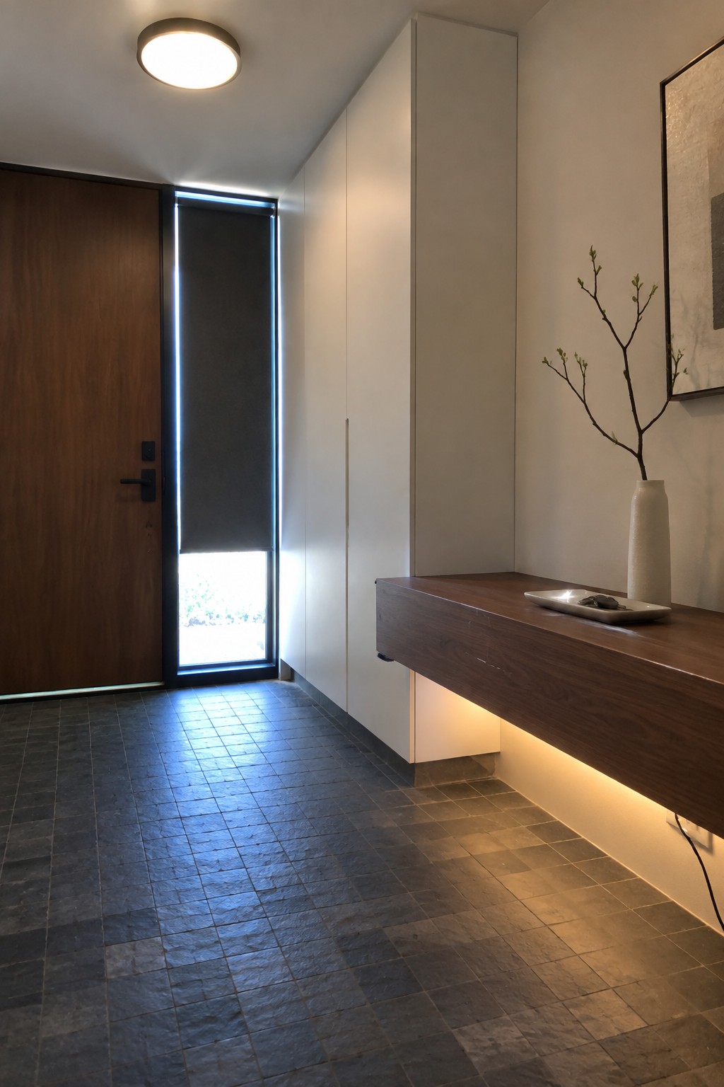A minimalist interior entryway with a wooden door featuring vertical backlighting, white built-in cabinets, a floating wood console holding a vase, and dark tiled flooring under a round ceiling light.