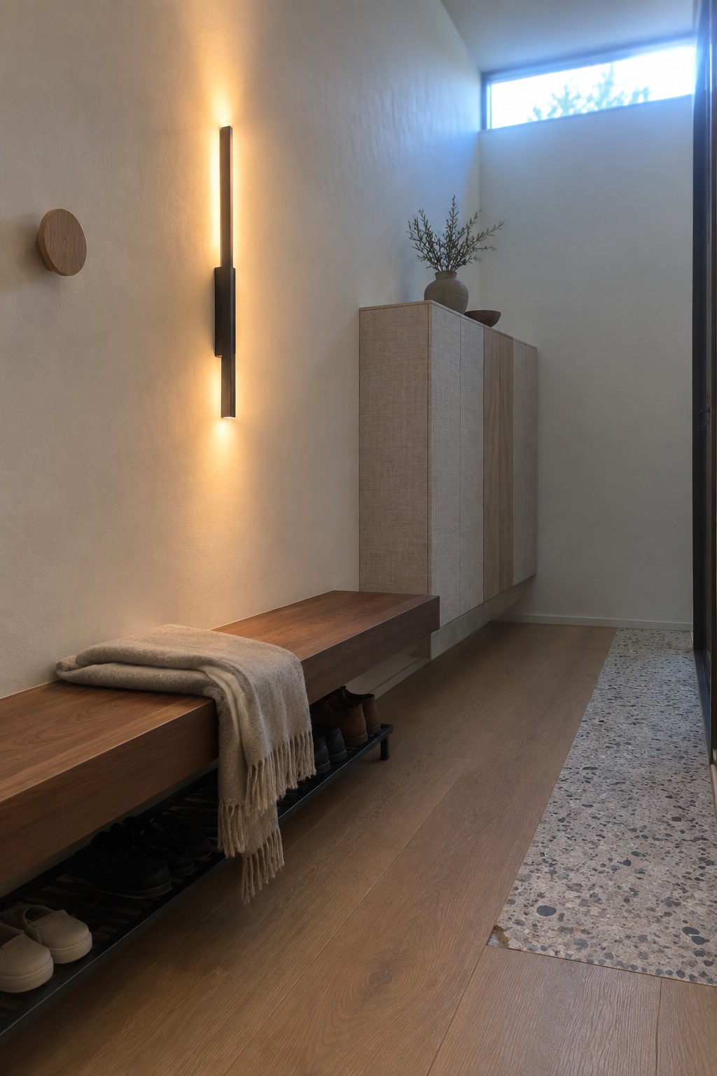A minimalist interior entryway with a tall vertical wall sconce above a floating wooden bench that has under-storage for shoes.