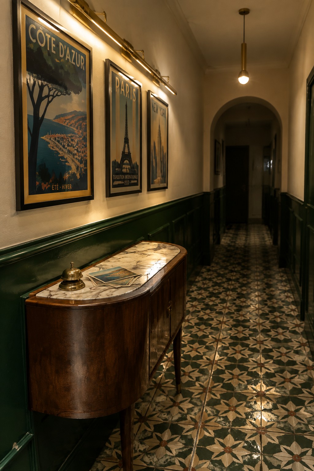 A narrow interior hallway with green wainscoting, three framed vintage travel posters lit by wall-mounted linear lights, and a curved wooden console table beneath them.