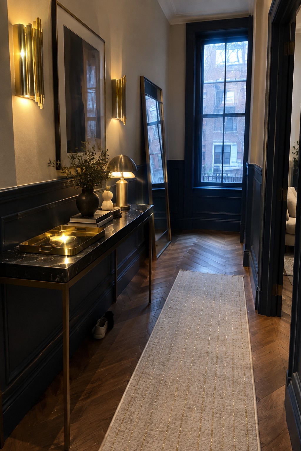 A narrow interior entry hallway with dark wainscoting, a marble-top console table, brass wall sconces next to framed art, and a tall mirror leaning against the wall.
