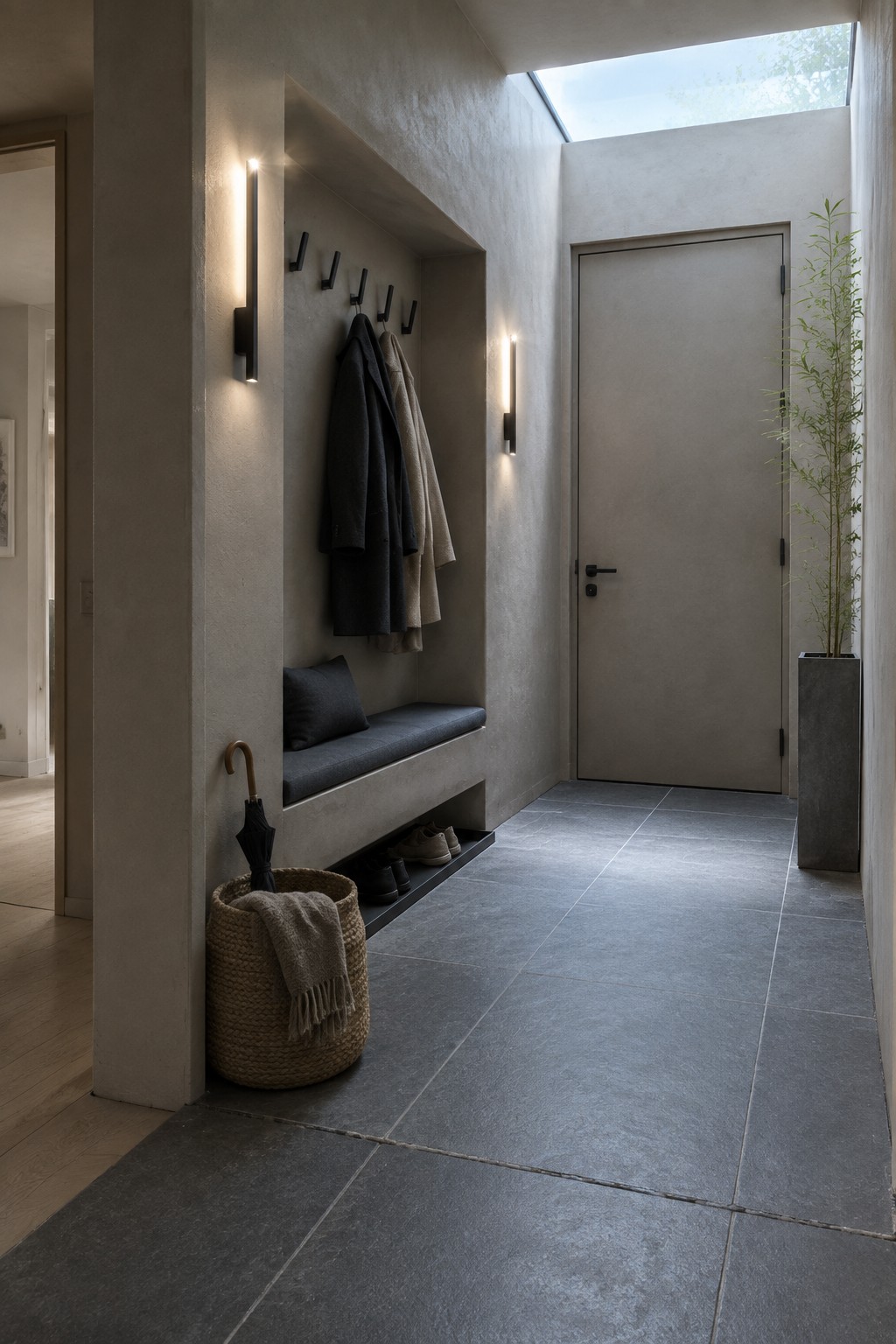 A narrow interior entryway with a recessed concrete bench, wall hooks holding coats, vertical wall sconces, and dark tile flooring.