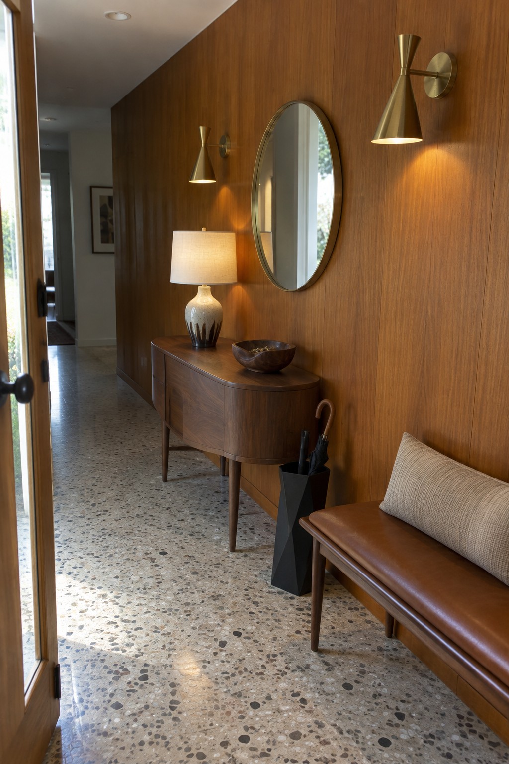 A narrow interior entry hallway with vertical wood paneling, two brass wall sconces, a round mirror, a wooden console table holding a lamp and bowl, a brown leather bench with cushion, and a black umbrella stand on speckled terrazzo flooring.
