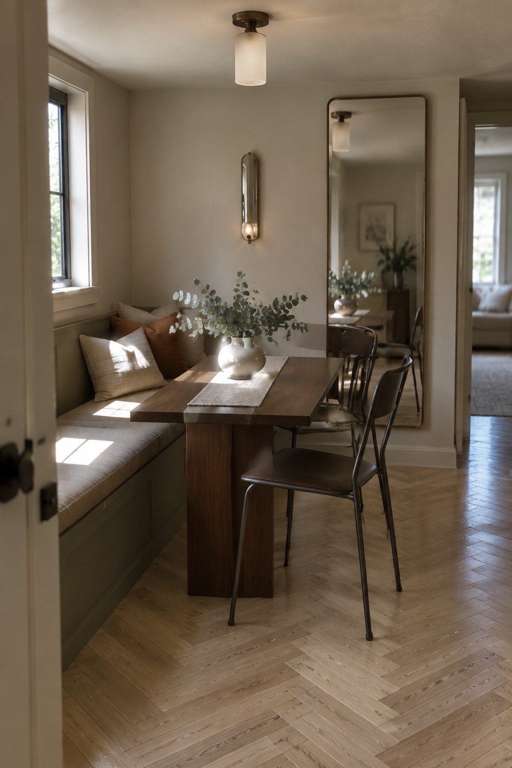 A dining nook with a wooden table, built-in bench with pillows, metal chairs, a vase of greenery, wall sconce, and ceiling light fixture.