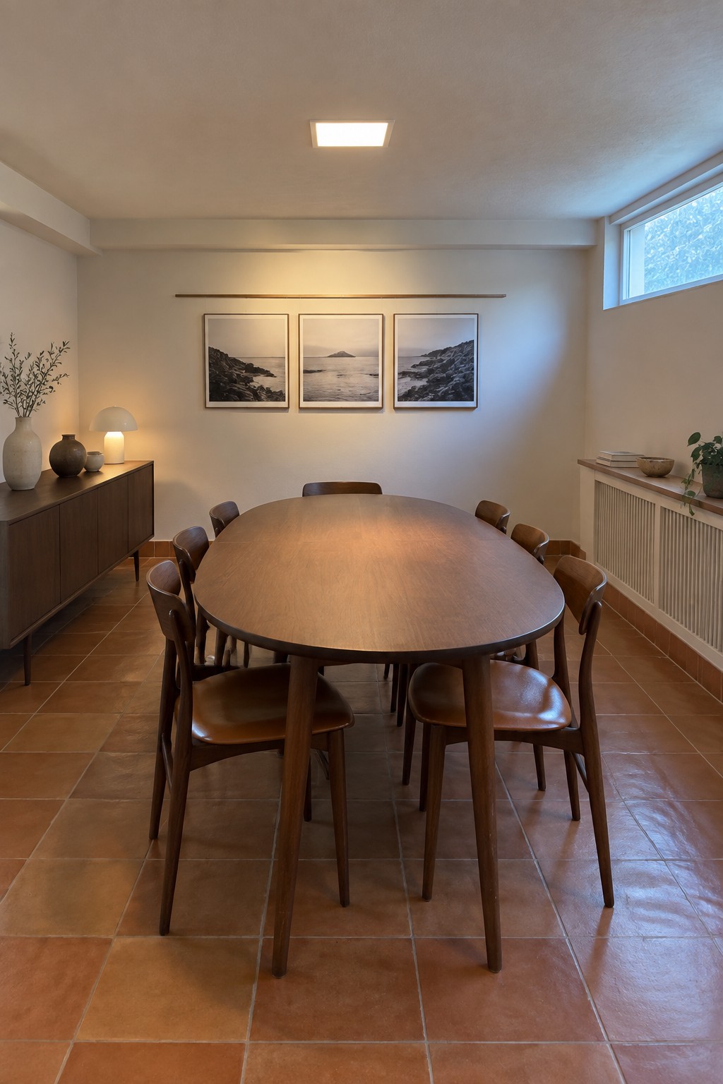 A dining room with an oval wooden table and eight chairs, three black and white photos on the wall, a wooden sideboard, and a square flush ceiling light.