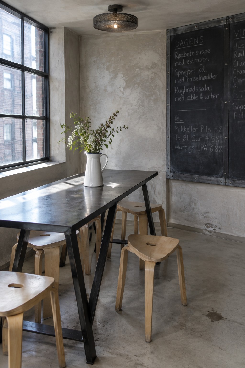 Industrial cafe with long metal table, wooden stools, chalkboard menu, and flower vase.