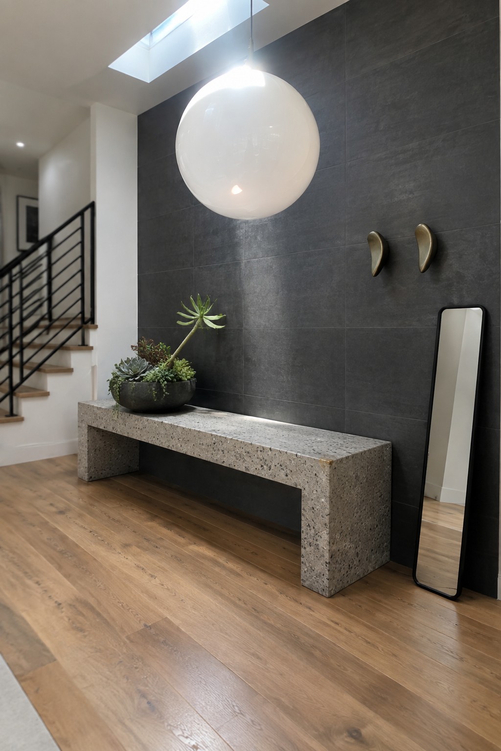 A foyer interior showing a large white spherical pendant light above a long concrete bench against a dark tiled wall with a tall mirror leaning nearby.