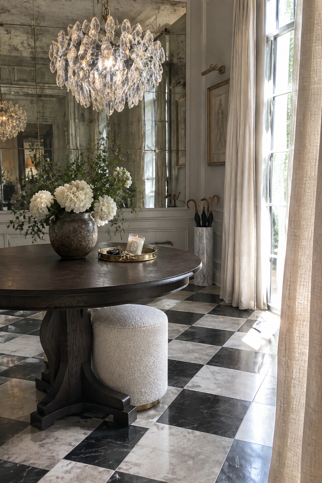 A large crystal chandelier hangs above a round dark wood table in a foyer with black and white marble checkered flooring and a tall mirror on the wall.