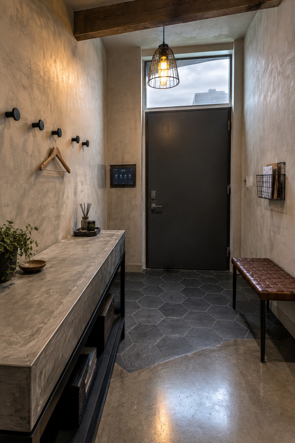A small modern foyer with a dark front door, concrete console table on the left, brown leather bench on the right, and a hanging wire cage pendant light centered above the entry.