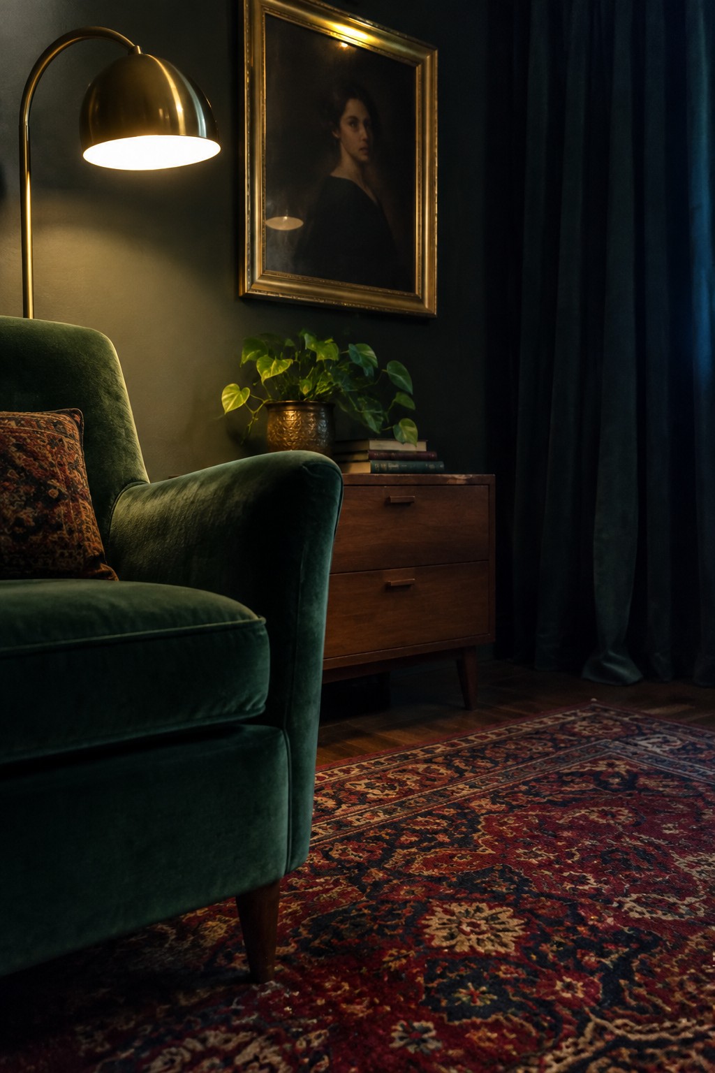 A green velvet armchair sits beside a wooden dresser with a brass floor lamp arching over the corner in a dark room with a red patterned rug.