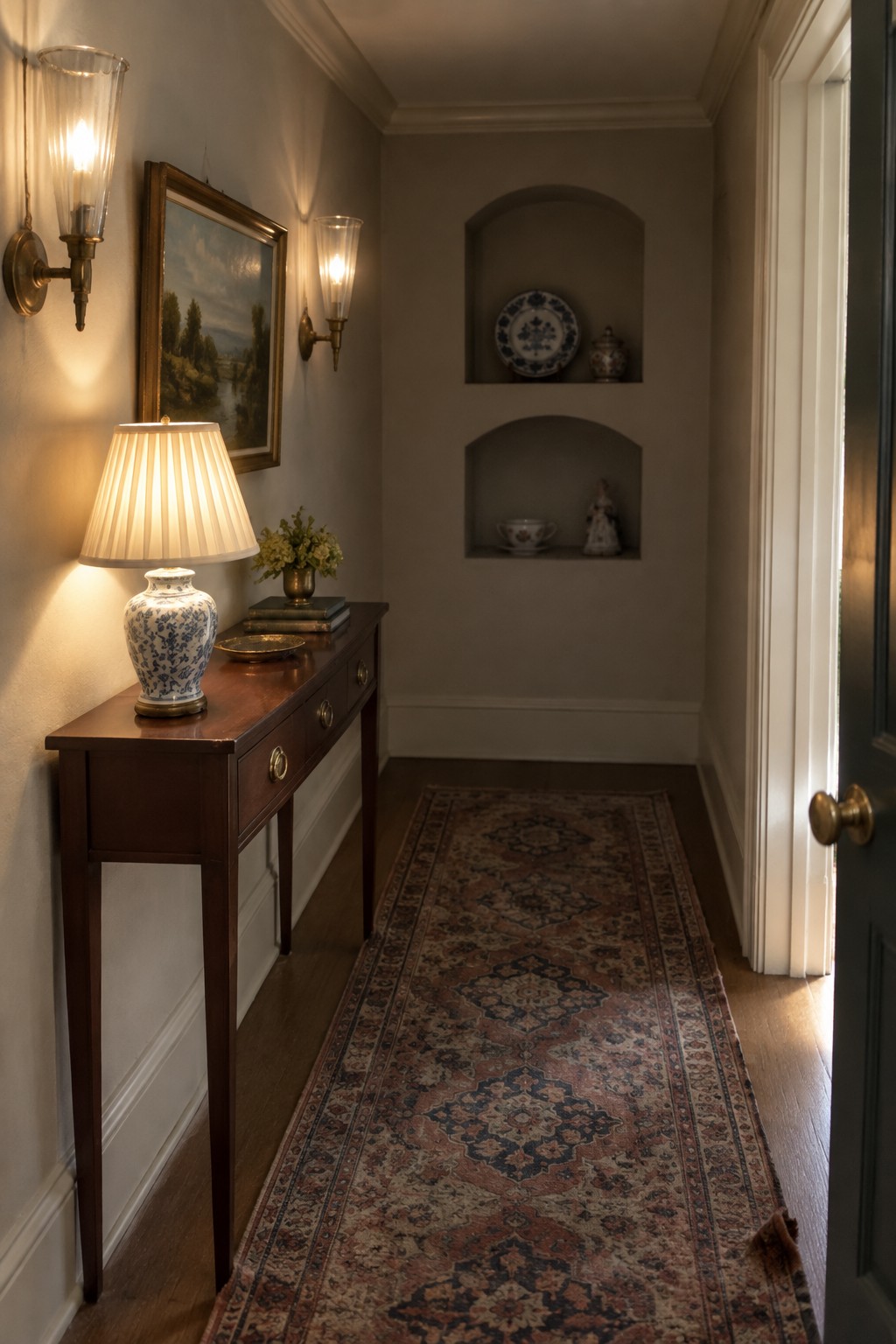 A narrow interior hallway with two wall sconces lighting a wooden console table below a framed painting and a long patterned runner on the floor.