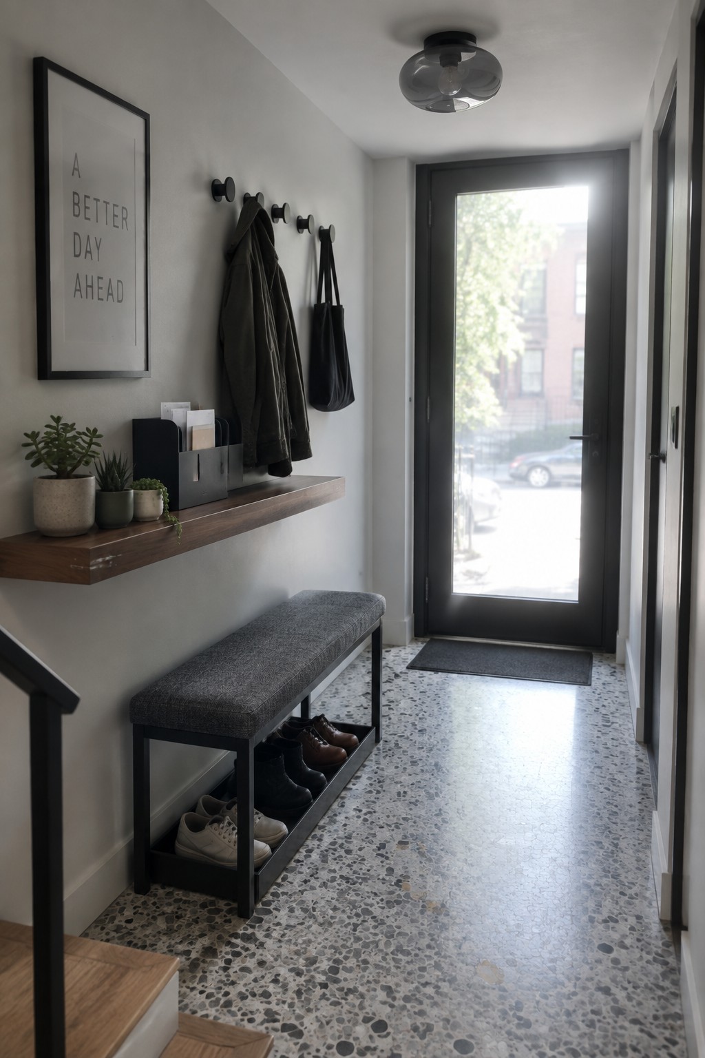A narrow interior foyer with a flush mount glass ceiling light, wooden floating shelf with plants and mail organizers, wall hooks holding a jacket and tote, gray upholstered bench with open shoe storage below, and black front door at the end.