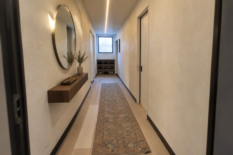 A narrow interior hallway with light wood floors, a long runner rug, a continuous linear ceiling light, and a round wall mirror on the right.