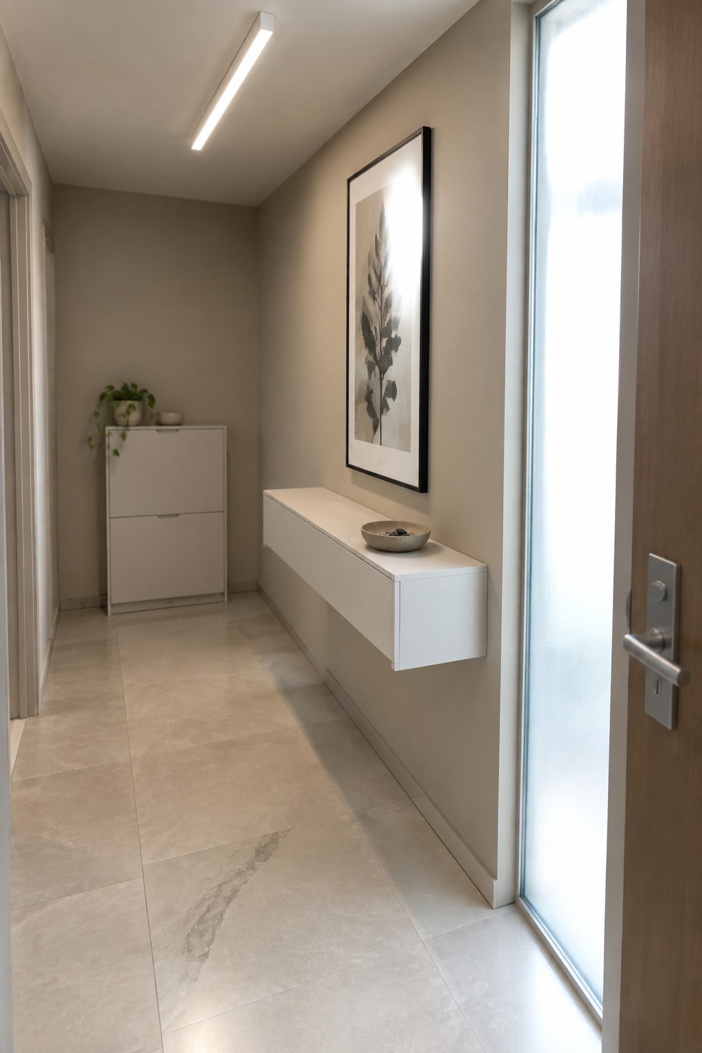 A narrow interior hallway with beige walls, large light tile flooring, a long linear ceiling light fixture, a floating white console table, and a framed botanical print on the wall.