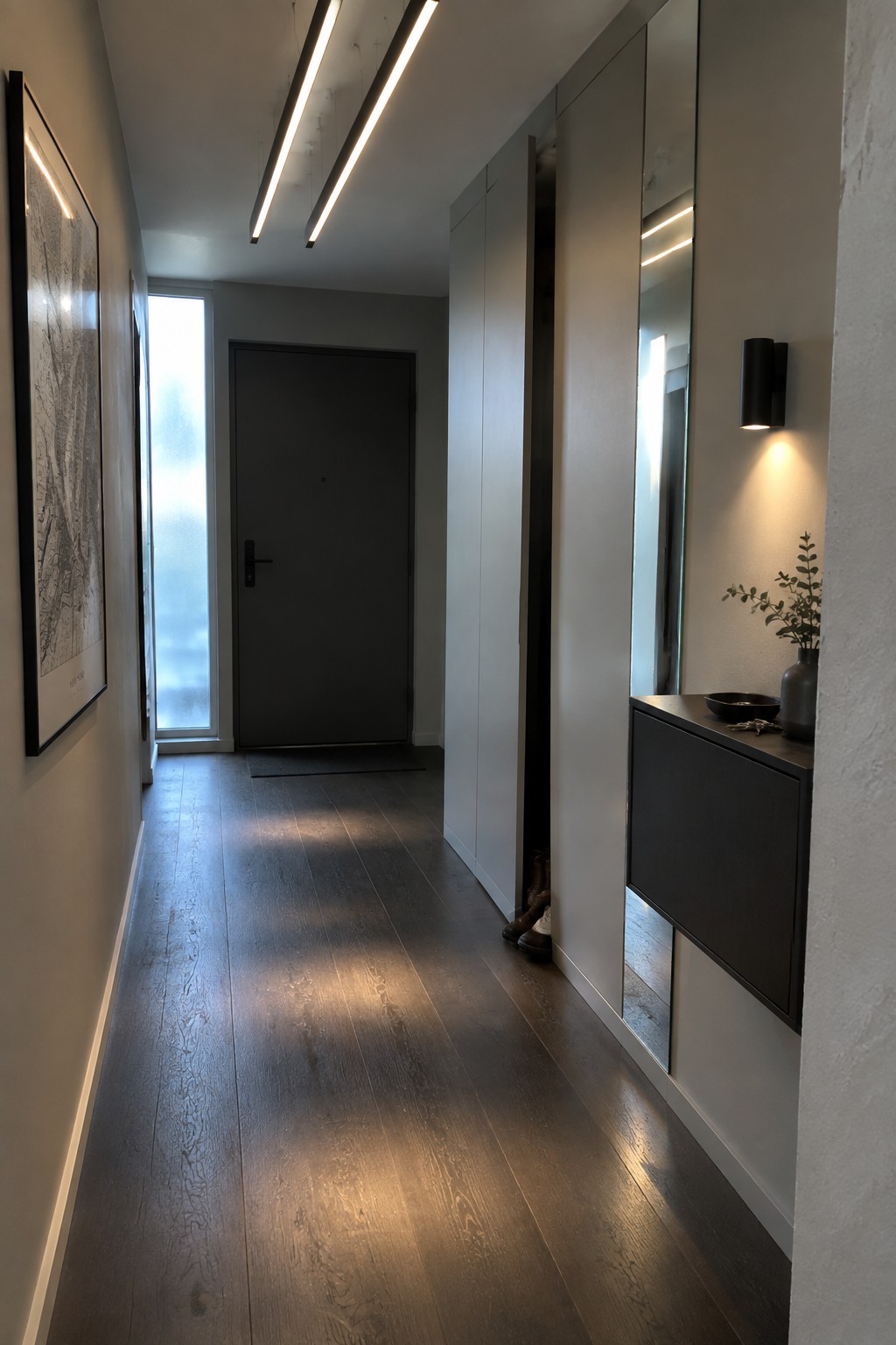 A narrow interior hallway with dark wood flooring, white walls, built-in cabinetry on the right, and two long linear light fixtures suspended from the ceiling.