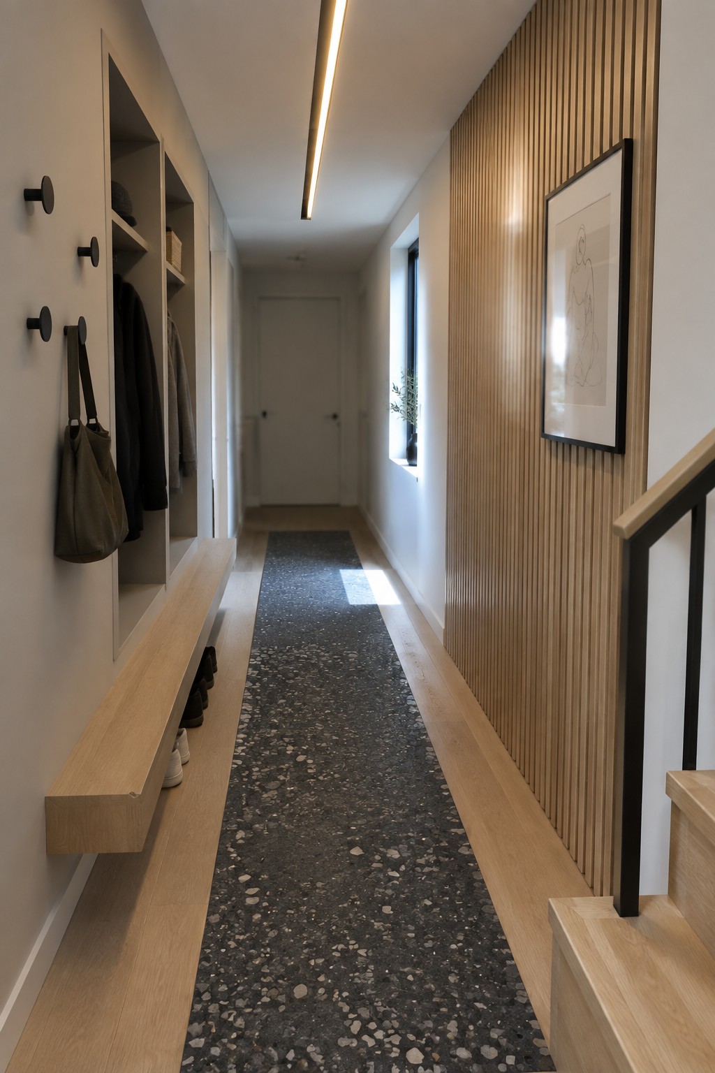 A narrow hallway with a long linear ceiling light fixture, light wood built-in storage and bench on the left, and a dark speckled runner rug on the floor.