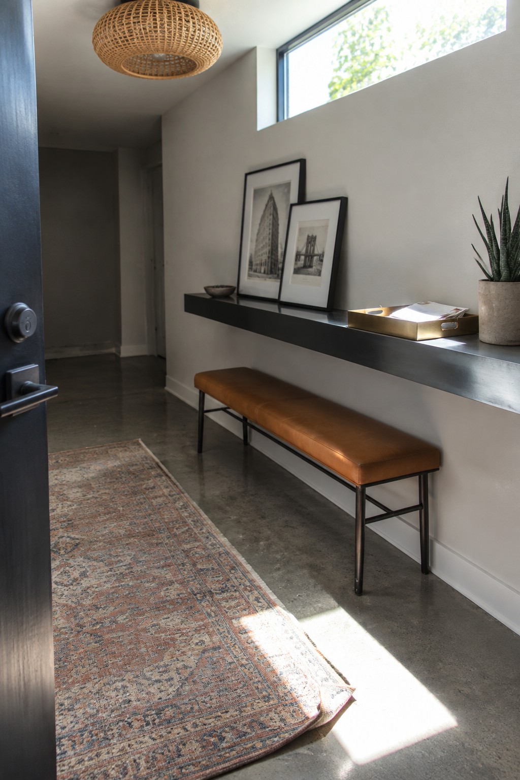 A woven rattan pendant light hangs from the ceiling in a minimalist entryway with a long bench, floating shelf, framed art, and a potted plant.